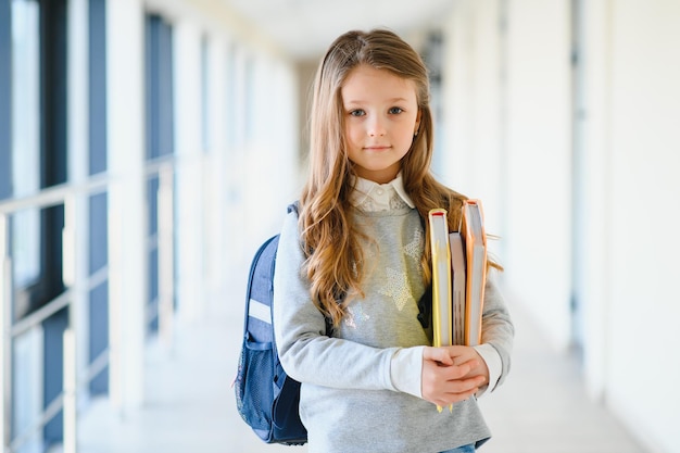 Vista frontal de la pequeña y hermosa niña de la escuela entre el pasillo de la escuela sosteniendo notas en las manos Niña divertida y feliz sonriendo a la cámara descansando después de las lecciones en la escuela primaria