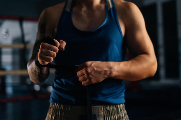 Vista frontal de primer plano de un boxeador irreconocible que envuelve cinta de boxeo negra alrededor de los brazos antes de pelear en un club deportivo con interior oscuro, en el fondo borroso del anillo. Luchador fuerte preparándose para usar guantes