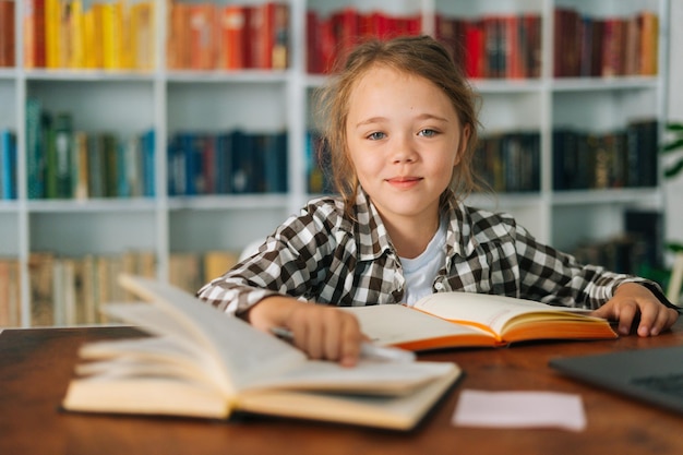 Vista frontal de primer plano de una linda alumna de la escuela infantil haciendo la tarea escribiendo en un cuaderno sentado en la mesa con un libro de papel, mirando la cámara. Retrato colegiala de niño elemental estudiando en casa.