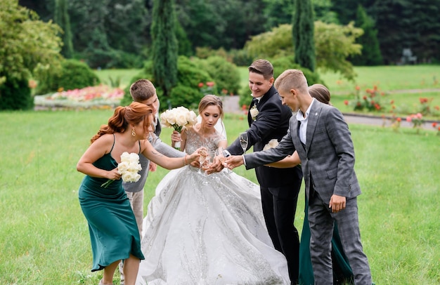 Vista frontal de recién casados con amigos sosteniendo copas de vino con bebidas de pie juntos y celebrando el día de la boda en un jardín increíble