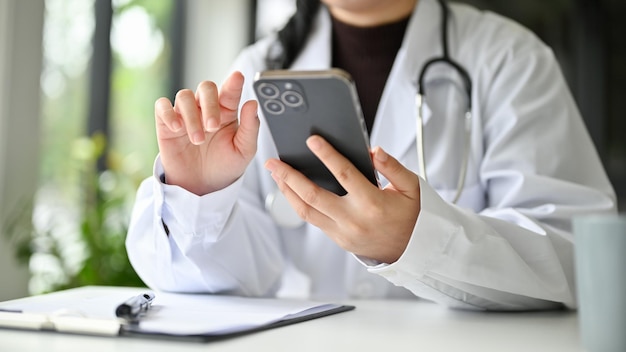 Vista frontal recortada de uma médica asiática profissional em vestido branco usando seu smartphone