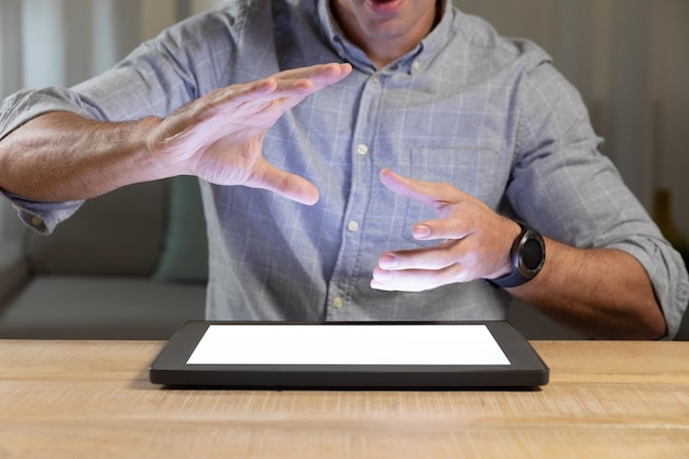 Vista frontal de la sección media de un joven caucásico sentado a la mesa usando una tableta en casa, con las manos levantadas sobre la pantalla iluminada, como si estuviera sosteniendo o tocando
