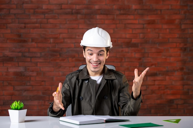 Vista frontal, sorrindo, engenheiro masculino, sentado atrás de seu local de trabalho e escrevendo notas, plano de documento, empreiteiro de propriedade, agenda, construtor trabalho corporativo