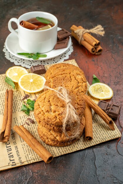 Vista frontal de una taza de té con limones amarillos frescos y galletas sobre un fondo oscuro, desayuno dulce de la mañana, té de frutas, ceremonia de color