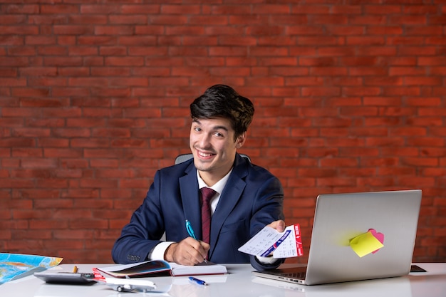 Vista frontal del trabajador masculino sentado detrás de su lugar de trabajo en traje dando par de boletos de avión trabajo ocupación proyecto en el extranjero trabajo turismo plan corporativo