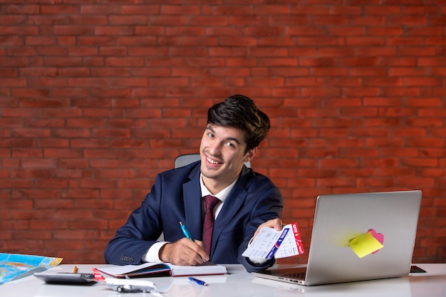 Vista frontal del trabajador masculino sentado detrás de su lugar de trabajo en traje sosteniendo un par de boletos de avión planificar trabajo proyecto de trabajo turismo corporativo en el extranjero