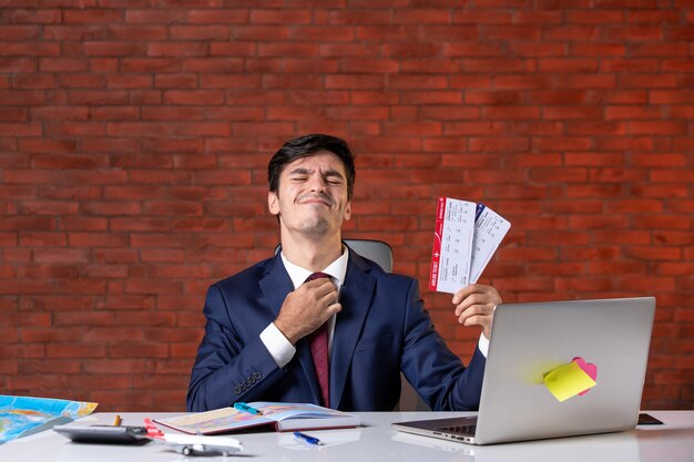 Vista frontal del trabajador masculino sentado detrás de su lugar de trabajo en traje sosteniendo un par de boletos de avión trabajo en el extranjero ocupación corporativa plan de proyecto de trabajo
