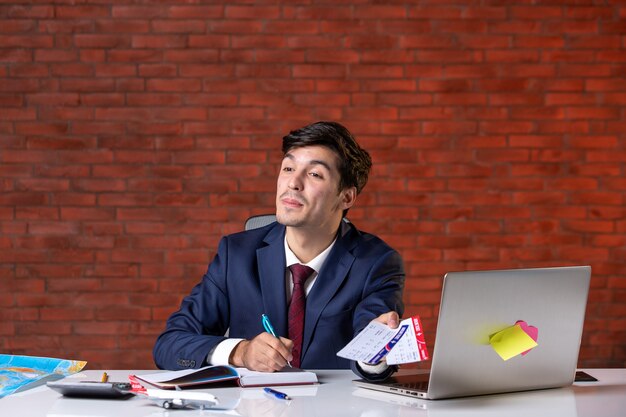 Vista frontal del trabajador masculino sentado detrás de su lugar de trabajo en traje sosteniendo un par de boletos de avión trabajo ocupación proyecto en el extranjero trabajo turismo plan corporativo