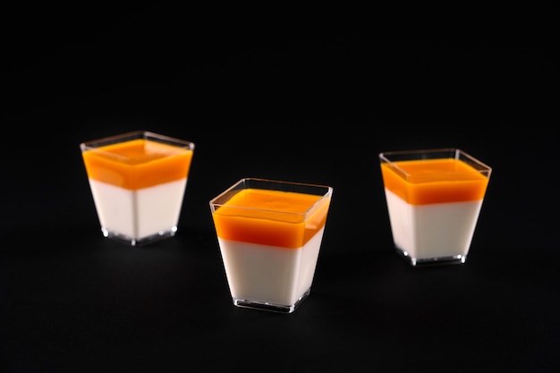 Vista frontal de tres vasos cuadrados pequeños con panna cotta lechoso decorado con una cobertura de color naranja brillante. Postre delicioso dulce aislado sobre fondo negro de estudio. Concepto de comida.