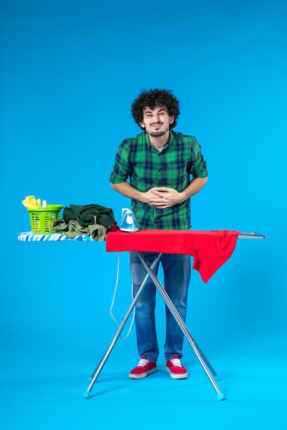 Vista frontal del varón joven con tabla de planchar con dolor de estómago sobre fondo azul lavadora limpia quehaceres domésticos color humano de la casa