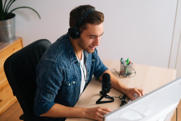 Vista de gran angular de un bloguero de audio positivo que usa audífonos sentado en el escritorio con un micrófono profesional y una computadora, transmitiendo podcast en el estudio de la oficina en casa con un interior moderno.