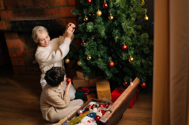 Vista de gran angular de la joven madre sonriente y su hijo decorando con coloridas bolas de árbol de Navidad sentados en el suelo cerca de la chimenea