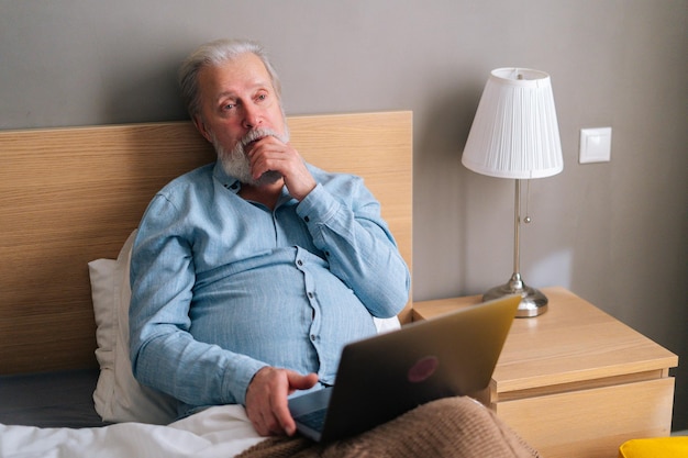 Vista de gran angular de pensar en un hombre mayor de pelo gris con barba usando una computadora portátil sentada en la cama en casa