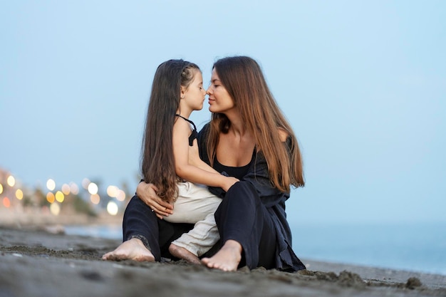 vista de la hermosa madre joven con la hija pequeña abrazada sentada en la playa con los ojos cerrados