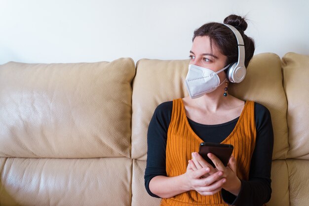 Vista horizontal da máscara protetora vestindo mulher doente, ouvindo música em casa. fique em casa. doença do vírus pandêmico covid 19. conceito de saúde.