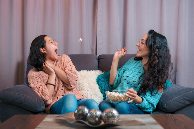 Vista horizontal de una madre y su hija viendo una película en casa juegan tirando palomitas de maíz para atraparlas con la boca hacen expresiones muy divertidas