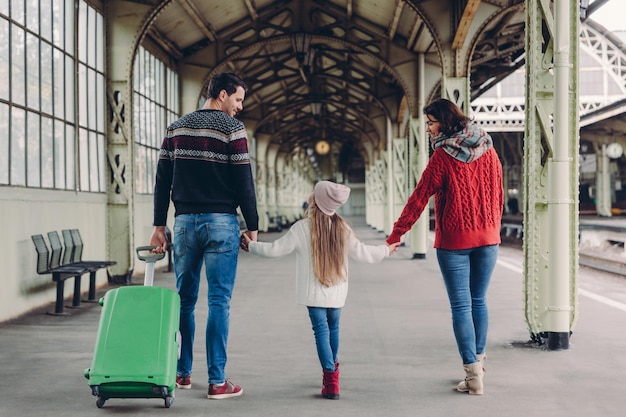 La vista horizontal de los padres cariñosos y su hija mantienen las manos juntas van a hacer un viaje al extranjero posan en la estación de tren llevan una bolsa verde tienen miradas felices comunican Viaje familiar