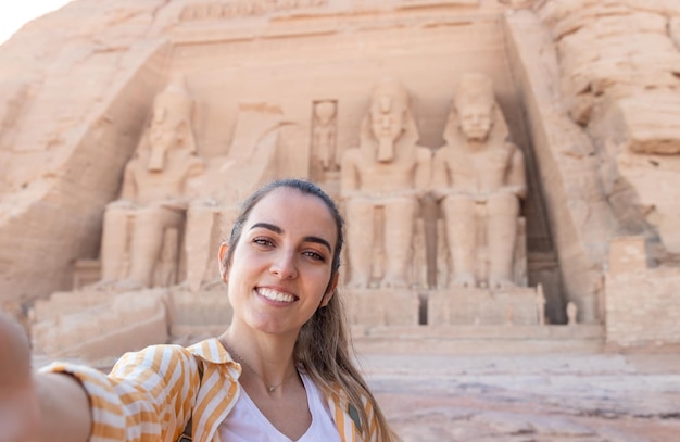 Vista horizontal de selfie de una joven y hermosa turista sonriente con el templo de Abu Simbel en el fondo