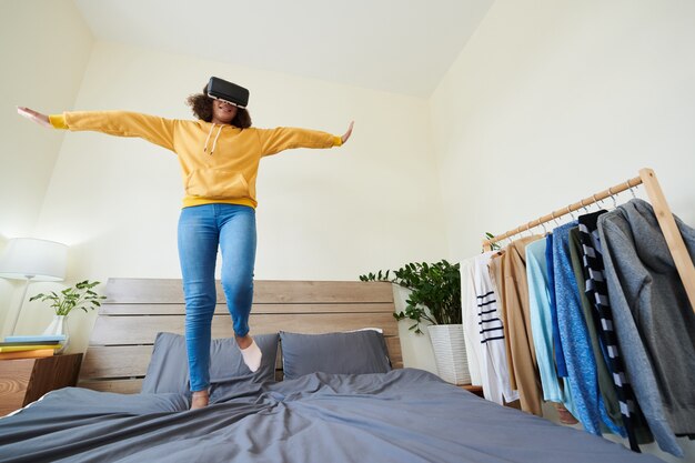 Vista inferior de la alegre niña de raza mixta despreocupada en gafas de realidad virtual saltando con los brazos extendidos en la cama mientras juega el juego D