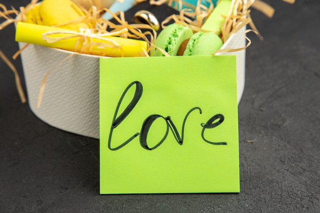 vista inferior caixa em forma de coração com macarons de anel enrolados em notas auto-adesivas amor escrito em nota auto-adesiva em fundo escuro