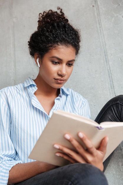 Vista inferior de uma jovem africana vestindo uma camisa, ouvindo música com fones de ouvido sem fio, enquanto está sentada na parede em casa e lendo um livro