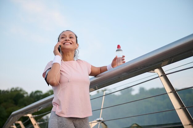 Vista inferior de uma jovem hispânica alegre em camiseta rosa falando no celular enquanto descansava em uma ponte da cidade após o treino de manhã cedo ao amanhecer. Conceito de estilo de vida ativo e saudável