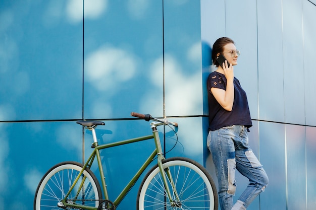 Vista inferior de la hermosa mujer elegante con gafas de sol conversando por teléfono móvil, de pie con bicicleta en la esquina del edificio moderno