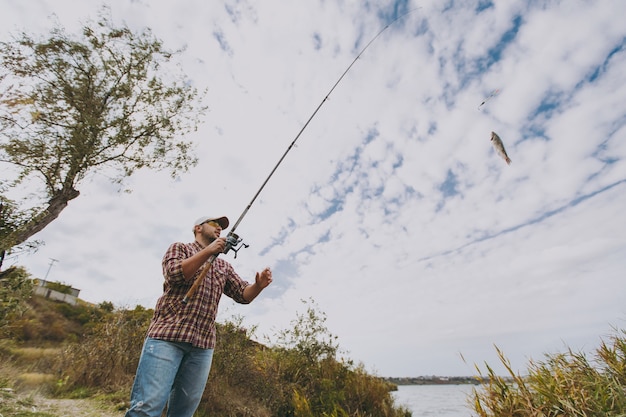 Vista inferior Joven sin afeitar con camisa a cuadros, gorra y gafas de sol saca una caña de pescar con pescado capturado en un lago desde la orilla cerca de arbustos y cañas. Estilo de vida, recreación, concepto de ocio.