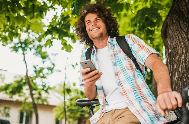 Vista inferior de un joven estudiante feliz sonriendo con el pelo rizado usando camisa con mochila sentado en la bicicleta en la calle relajando mensajes de texto en teléfonos inteligentes Personas y educación
