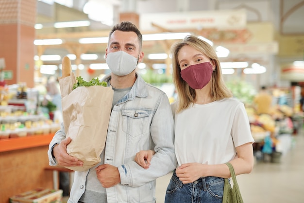 Vista inferior de una pareja joven con máscaras faciales caminando juntos sobre el mercado de agricultores durante el coronavirus y comprando alimentos