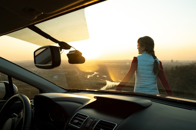 Vista desde el interior de una mujer joven de pie cerca de su coche disfrutando de la cálida vista del atardecer.