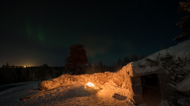 Vista invernal de fuego cerca de la cabaña y la aurora boreal en el oscuro cielo nocturno, Finlandia