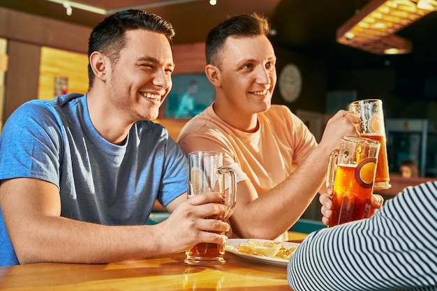 Vista desde el lado de dos hombres felices descansando con amigos en el bar, bebiendo cerveza y riendo juntos. Hombre alegre mirándose, bromeando y hablando los fines de semana. Concepto de ocio y diversión.