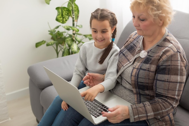 Vista lateral de una abuela y una nieta usando una computadora portátil en la sala de estar en casa