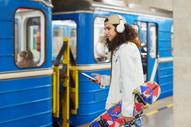 Vista lateral de un adolescente moderno con auriculares parados frente al tren subterráneo azul