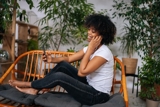 Vista lateral de una alegre joven afroamericana escuchando música desde un teléfono móvil eligiendo la próxima canción sentada en el sofá en casa