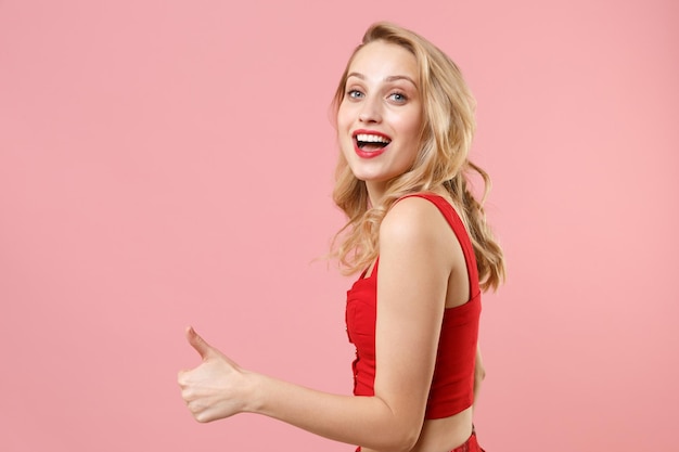Vista lateral de una alegre joven mujer rubia con ropa sexy roja posando aislada en un retrato de estudio de fondo rosa pastel. Concepto de estilo de vida de las emociones de las personas. Simulacros de espacio de copia. Mostrando el pulgar hacia arriba.