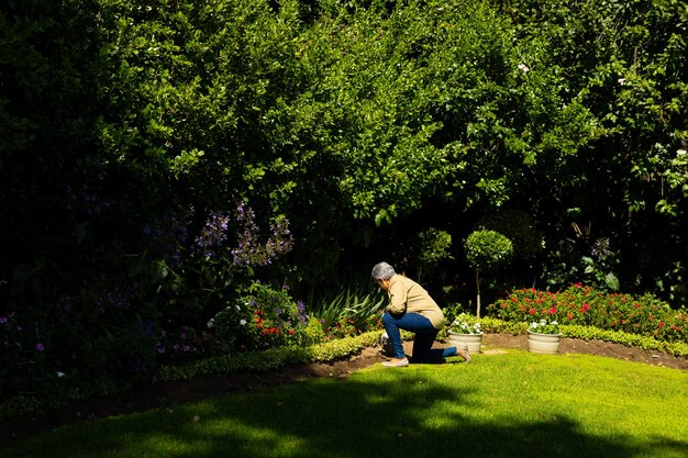 Vista lateral de una anciana birracial con jardinería de pelo corto mientras se arrodilla en un terreno cubierto de hierba en el patio