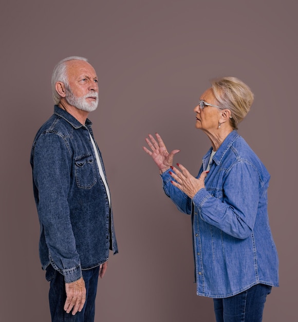 Vista lateral de una anciana enojada y frustrada gesticulando y discutiendo con su esposo ignorándola y mirando hacia otro lado aislada en un fondo de color