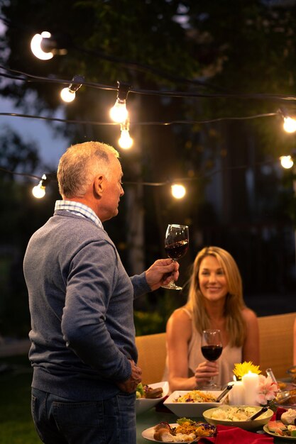 Vista lateral de un anciano caucásico de pie y haciendo un brindis con una copa de vino tinto, durante una comida de celebración familiar afuera, su hija adulta sentada en la mesa y mirándolo