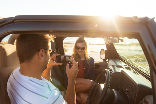 Vista lateral de ángulo alto de un hombre caucásico con su novia sonriendo dentro de un coche abierto, tomando una foto de ella