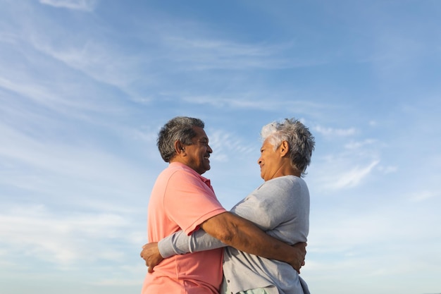 Vista lateral en ángulo bajo de una sonriente pareja de ancianos birraciales mirando mientras se abrazan contra el cielo