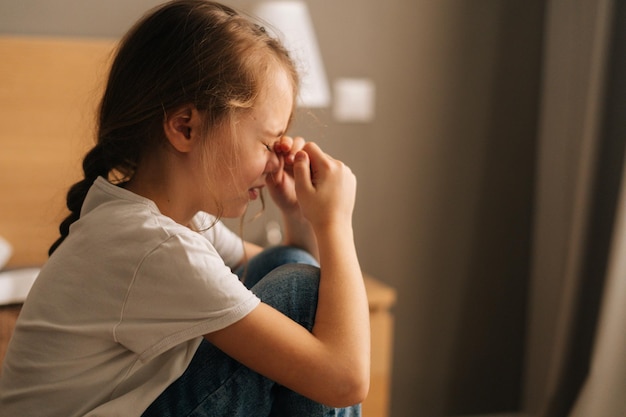 Foto vista lateral aproximada de uma garotinha chateada chorando sentada sozinha na cama no quarto conceito de depressão infantil