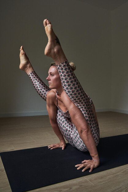 Vista lateral de la atleta femenina enfocada en el equilibrio de ropa activa en Tittibhasana en la alfombra mientras hace yoga en la habitación y estira las piernas