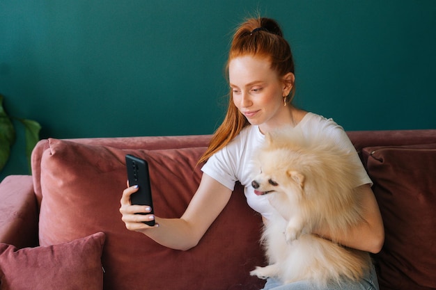 Vista lateral de una atractiva joven pelirroja abrazando a un pequeño y blanco perro mascota Spitz y tomándose selfie usando un teléfono móvil sentado en un cómodo sofá, en una acogedora sala de estar luminosa con un interior moderno.