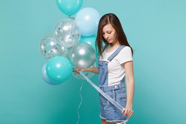 Vista lateral de una atractiva joven vestida de denim mirando hacia abajo celebrando sosteniendo coloridos globos de aire aislados en un fondo azul turquesa de la pared. Fiesta de cumpleaños, concepto de emociones de la gente.