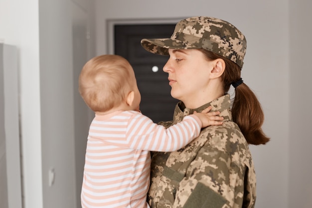 Vista lateral de la atractiva madre militar vestida con uniforme de camuflaje y sombrero, sosteniendo a su hijo y mirando a su hija, tan esperado encuentro mujer soldado con su hijo.