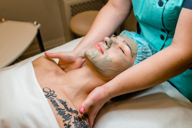 Vista lateral baja de manos con guantes blancos haciendo masaje manual de ojos faciales La mujer joven tiene una máscara de crema blanca en la cara Procedimientos de spa de belleza