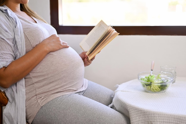 Vista lateral y captura recortada de una mujer asiática embarazada leyendo un libro en la mesa del comedor