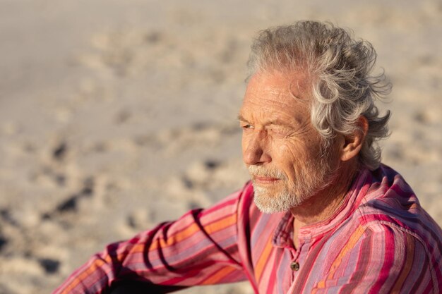 Vista lateral de cerca de un anciano caucásico en la playa bajo el sol, sentado en la arena, sonriendo y mirando al mar